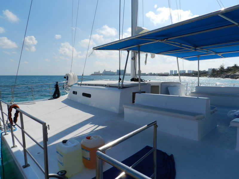 Aft deck view of Catamaran Samba 3 at sea with blue canopy shade, Cancun hotel zone visible in background, large group sailing charter