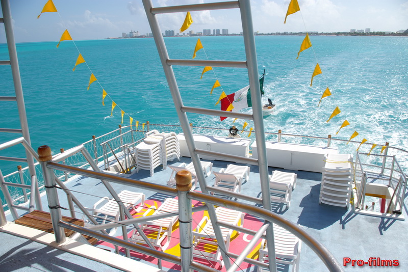 Upper sun deck of the Dancer catamaran lined with white lounge chairs and yellow pennant flags overlooking turquoise water and the Cancun skyline