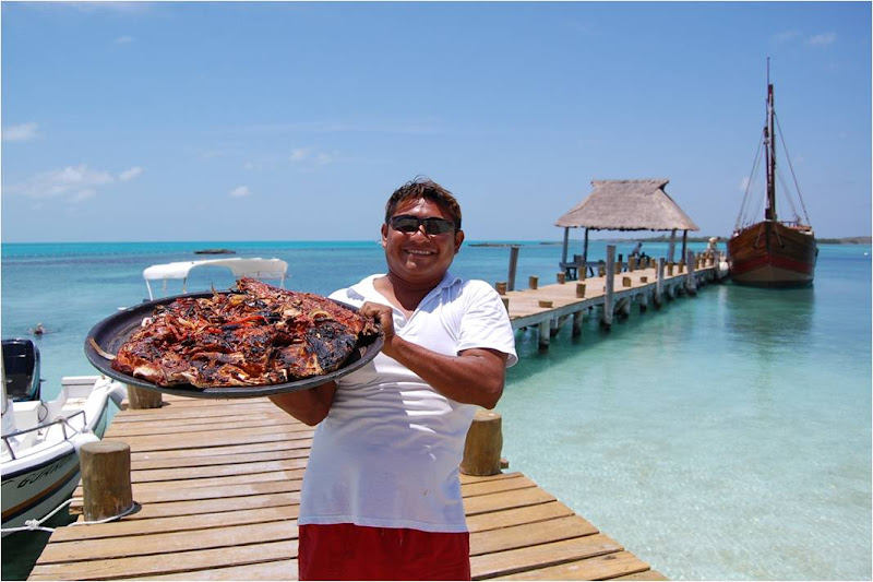 Smiling crew member holding a large tray of freshly grilled fish on the pier with the caravel and turquoise sea behind at Isla Contoy