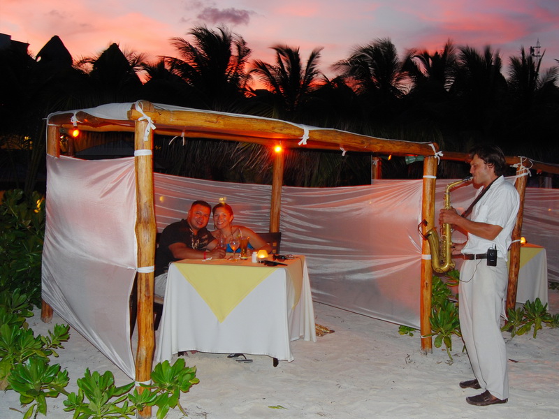 Couple dining in a draped beach cabana while a saxophonist plays at sunset on Maroma beach, romantic Riviera Maya catamaran dinner cruise