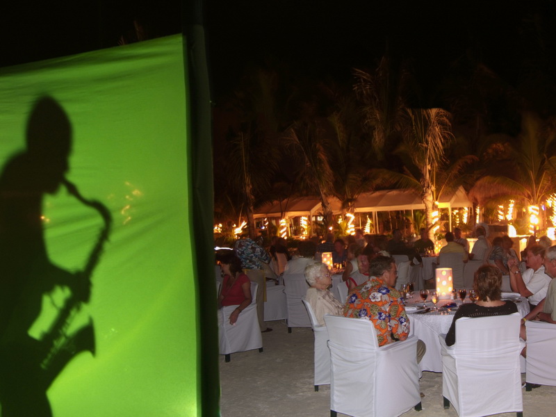 Beachfront candle-lit dinner tables with guests and a saxophonist silhouette glowing at night on Maroma beach, Playa del Carmen Riviera Maya