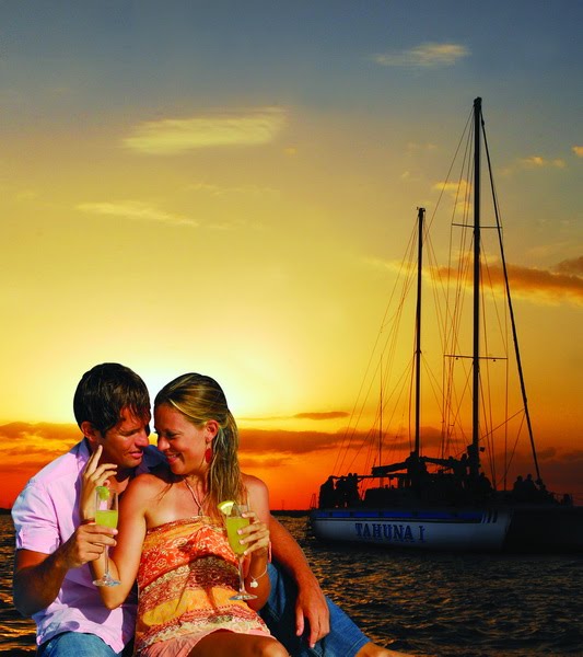 Couple toasting cocktails as the Tahuna sailing catamaran is silhouetted against a golden Caribbean sunset off Maroma Playa del Carmen